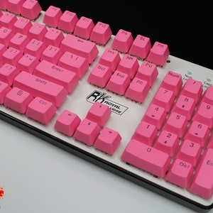 Pu&ograve; includere: Una tastiera meccanica con tasti rosa acceso. La tastiera ha una base bianca con finiture nere e presenta tasti standard, inclusi tasti funzione, un tastierino numerico e tasti freccia. Il marchio "RK ROYAL KLUDGE" &egrave; visibile.