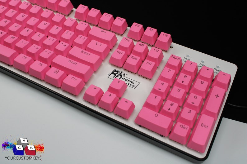 Pu&ograve; includere: Una tastiera meccanica con tasti rosa acceso. La tastiera ha una base bianca con finiture nere e presenta tasti standard, inclusi tasti funzione, un tastierino numerico e tasti freccia. Il marchio "RK ROYAL KLUDGE" &egrave; visibile.