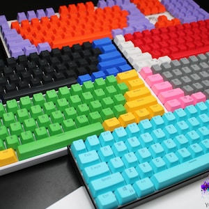 May include: A collection of colorful mechanical keyboards with various keycap colors, including blue, green, orange, and pink. The keyboards are arranged in a visually appealing display, showcasing different color schemes and layouts. The image includes the text "YOURCUSTOMKEYS".