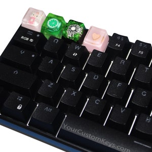 Può includere: Tastiera meccanica nera con tasti personalizzati. I tasti includono un coniglietto rosa, una fetta di lime verde, un design floreale verde e un cuore rosa. La tastiera ha il testo "YourCustomKeys.com" in basso.