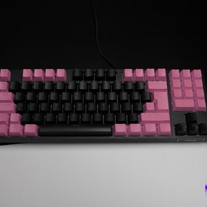 May include: A black mechanical keyboard with a mix of black and pink keycaps. The keyboard has a wired connection and is set against a black and white background. The brand name is visible on the bottom right.