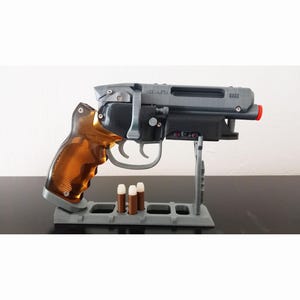 May include: A grey and black toy gun with a copper-coloured handle and a red tip. The gun is resting on a grey stand with three brown bullets. The text "MADE IN ETA" and "8223" are visible on the gun.