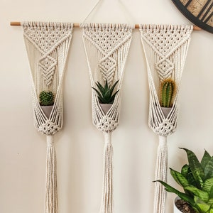Puede incluir: Tres colgadores de plantas de macram&eacute; con peque&ntilde;as suculentas y cactus en macetas. Los colgadores tejidos de color crema est&aacute;n suspendidos de un pasador de madera. Cada colgador tiene una borla. Fondo neutro.