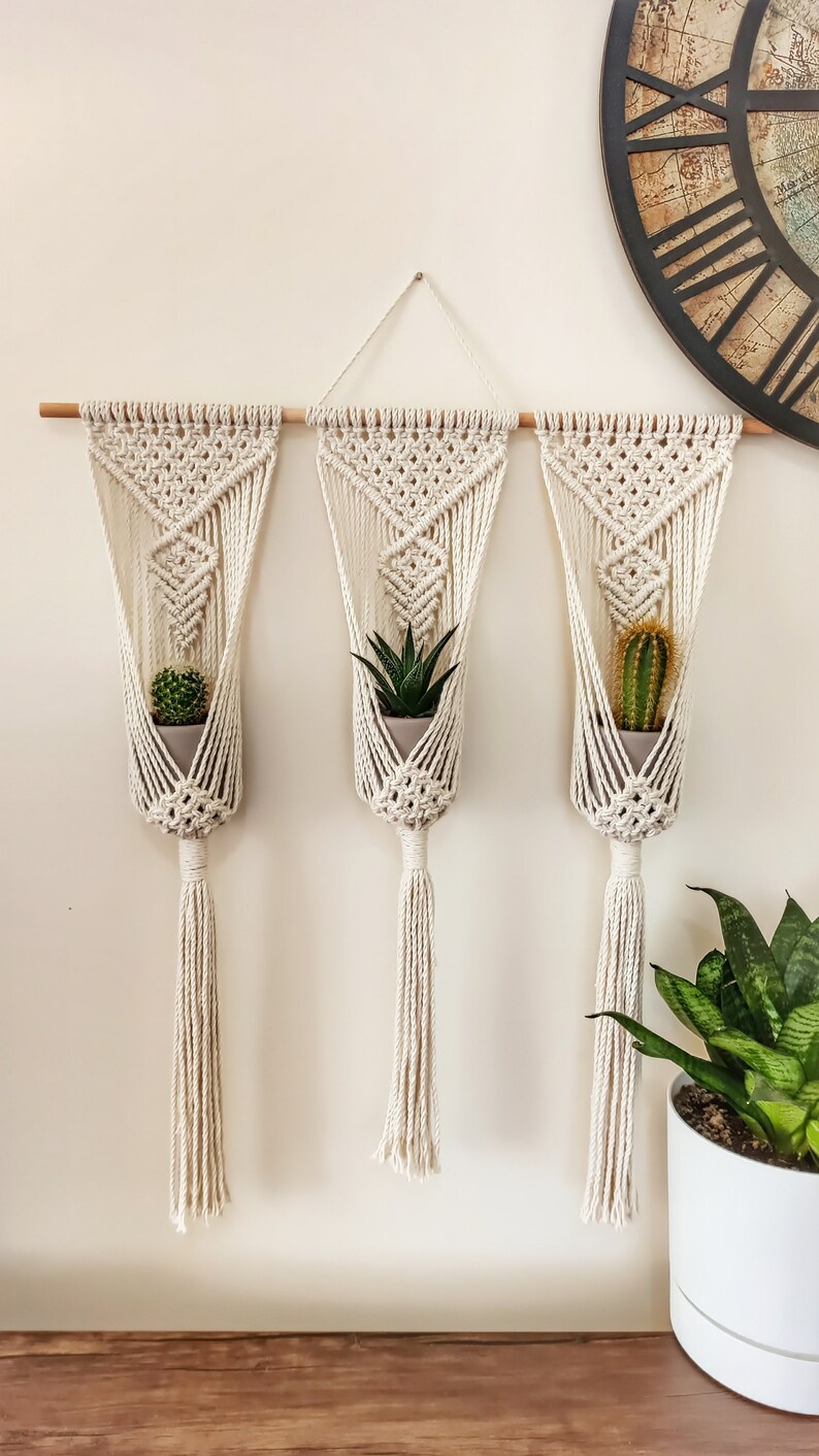 Puede incluir: Tres colgadores de plantas de macram&eacute; con peque&ntilde;as suculentas y cactus en macetas. Los colgadores tejidos de color crema est&aacute;n suspendidos de un pasador de madera. Cada colgador tiene una borla. Fondo neutro.