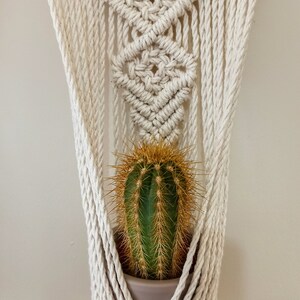 Puede incluir: Un colgador de plantas de macram&eacute; hecho a mano en algod&oacute;n blanco roto natural sostiene un cactus verde con espinas doradas en una maceta gris. El macram&eacute; presenta intrincados patrones de nudos y un dise&ntilde;o decorativo.