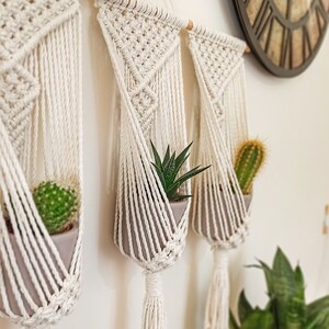 Puede incluir: Soportes de macram&eacute; color crema con macetas grises, cada uno con una suculenta o cactus. El macram&eacute; est&aacute; suspendido de un pasador de madera. Un reloj de pared grande y otras plantas est&aacute;n en el fondo.