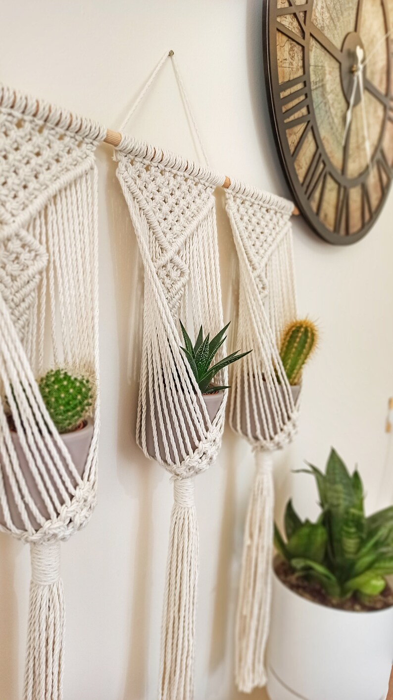Puede incluir: Soportes de macram&eacute; color crema con macetas grises, cada uno con una suculenta o cactus. El macram&eacute; est&aacute; suspendido de un pasador de madera. Un reloj de pared grande y otras plantas est&aacute;n en el fondo.