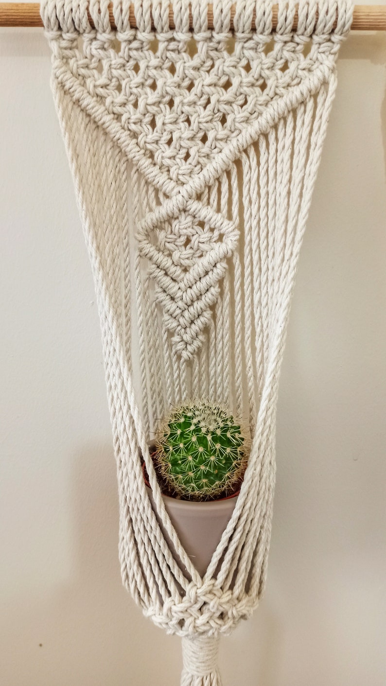 Puede incluir: Colgador de plantas de macram&eacute; color crema con dise&ntilde;o geom&eacute;trico, sosteniendo un peque&ntilde;o cactus verde en una maceta gris. El macram&eacute; est&aacute; suspendido de una varilla de madera, creando una est&eacute;tica decorativa y natural. El cactus mide unos 10 cm de di&aacute;metro.