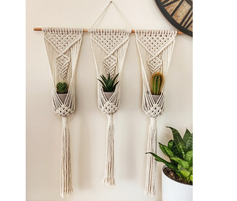 Puede incluir: Tres colgadores de plantas de macram&eacute; blanquecino suspendidos de una barra de madera. Cada colgador sostiene una peque&ntilde;a planta en maceta: un cactus, una suculenta y otro cactus. Los colgadores tienen un dise&ntilde;o geom&eacute;trico y largas borlas.