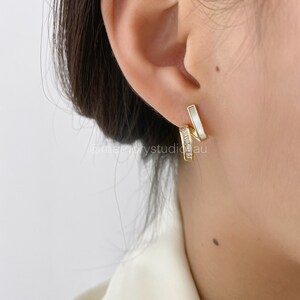 May include: Gold-toned earrings with a modern design. One section features a row of small, clear crystals, while the other has a white, rectangular inlay. The earrings are worn on an ear, showcasing their elegant style.