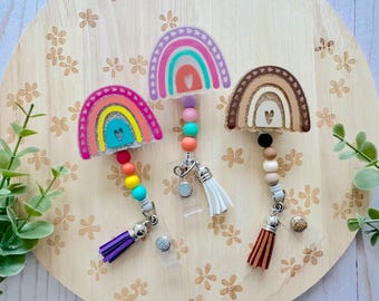 May include: Three rainbow-themed badge reels with colorful beaded accents and tassels. The reels feature rainbow designs in pink, purple, and brown. The background is a wooden surface with floral patterns.