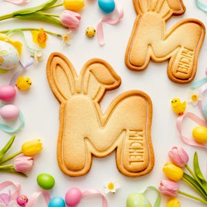 Può includere: Biscotti pasquali a forma di lettera "M" con orecchie da coniglio, decorati con il nome "MICHEL". I biscotti sono circondati da uova di Pasqua colorate, tulipani, nastri e piccole figurine di pulcini gialli, su sfondo bianco.