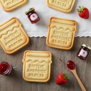 Peut inclure: Vue aérienne de biscuits en forme de pots de confiture, avec l'inscription "BAPTEME AMBROISE 05.07.26". De petits pots de confiture et des fraises fraîches sont disposés autour des biscuits sur une surface en bois.