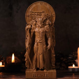 May include: A detailed wooden statue of a mythical figure with a horned head, intricate carvings, and a flowing robe. The statue is set on a base with the word "BUNE" carved into it. The background includes lit candles and pine cones.