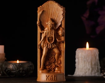 May include: Wooden statue of the Hindu goddess Kali, carved in detail. The figure is holding a sword and a severed head. The statue is set against a dark background with lit candles and the word "Kali" at the base.