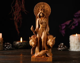 May include: A wooden statue of a woman with a halo, flanked by two wolves and a raven. The statue is light brown and stands on a base with the name "MELINOË". The background includes lit candles and dark, dried foliage.