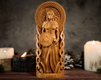 May include: A light brown wooden carving of a standing figure with long hair, wearing a textured garment and holding a circular shield with a triquetra symbol. The figure is framed by an intricate braided border and the name BRIGID is carved at the base.