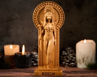 Puede incluir: Una talla de madera de color marrón claro de la deidad egipcia Sejmet, con cabeza de leona, un disco solar con una cobra y un cetro. La figura se alza sobre una base rectangular con el nombre SEKHMET grabado en mayúsculas en el frente.