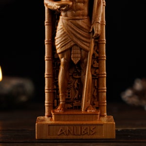 May include: A detailed wooden statue of Anubis, the Egyptian god of the afterlife, holding a staff and a bowl. The statue is carved with intricate details and hieroglyphs. The word "ANUBIS" is carved on the base.