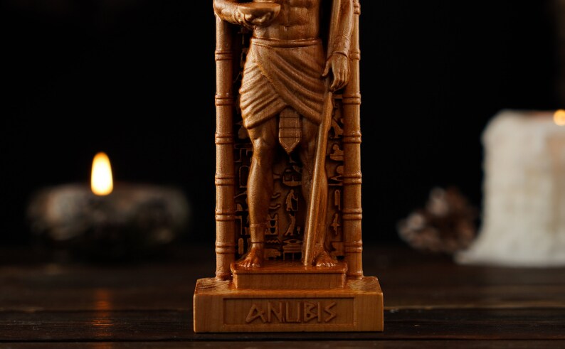 May include: A detailed wooden statue of Anubis, the Egyptian god of the afterlife, holding a staff and a bowl. The statue is carved with intricate details and hieroglyphs. The word "ANUBIS" is carved on the base.