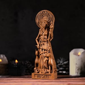 May include: A brown wooden statue of a figure holding a torch, surrounded by two dogs and skulls at the base. The figure has a large circular halo behind the head and the name HECATE is carved into the rectangular base in capital letters.