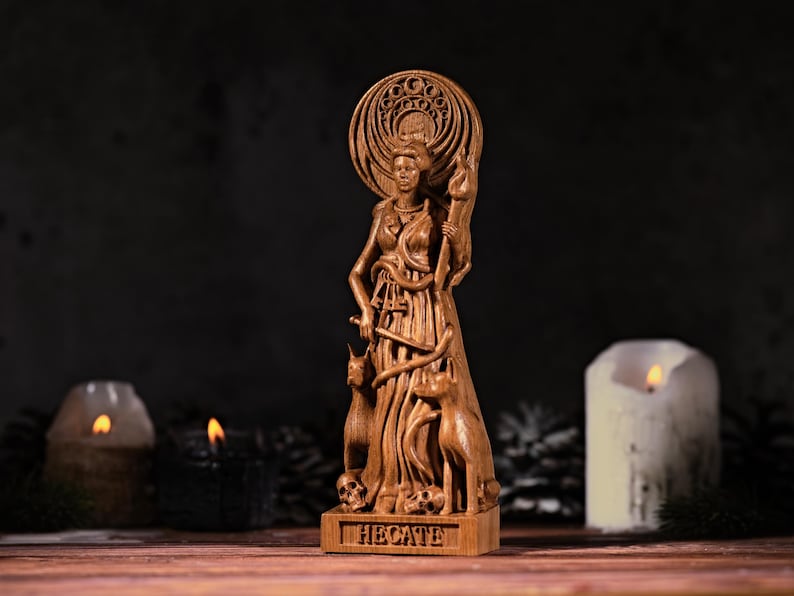 May include: A brown wooden statue of a figure holding a torch, surrounded by two dogs and skulls at the base. The figure has a large circular halo behind the head and the name HECATE is carved into the rectangular base in capital letters.