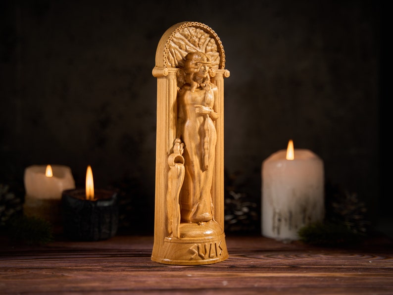 May include: A wooden statue of a woman with a tree behind her, standing in a carved archway. The statue is light brown and has the word "Sulis" carved at the bottom.