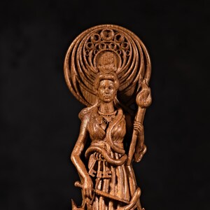 May include: A detailed wooden sculpture of a woman holding a torch and a sword. The figure has a halo-like design behind her head and is adorned with a snake. The statue is carved from a light brown wood.