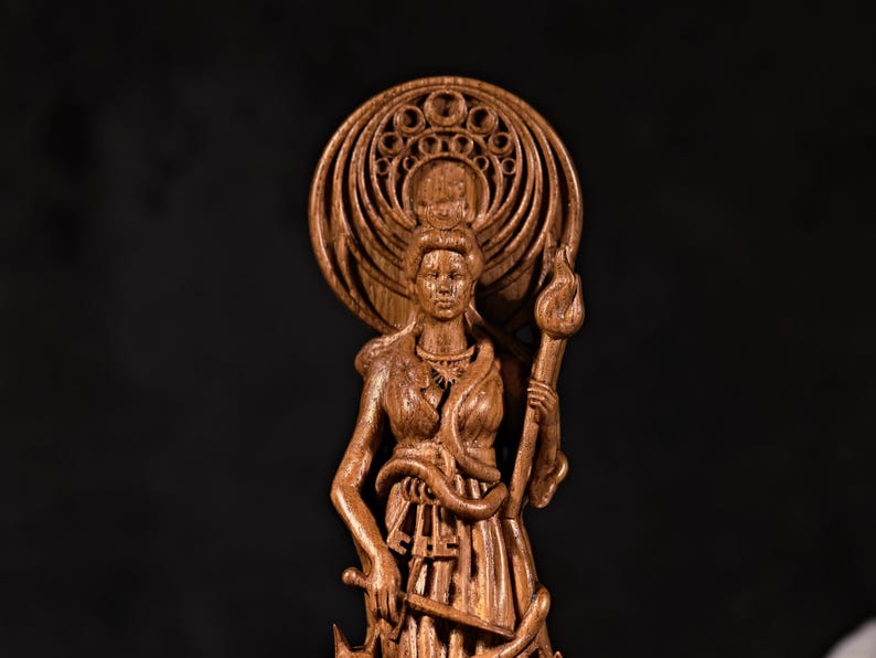 May include: A detailed wooden sculpture of a woman holding a torch and a sword. The figure has a halo-like design behind her head and is adorned with a snake. The statue is carved from a light brown wood.