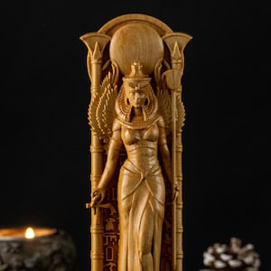 Op de afbeelding: Een gedetailleerde houten sculptuur van een Egyptische godin, met ingewikkelde details van haar kleding en hoofdtooi. De figuur staat tegen een donkere achtergrond, wat de natuurlijke houttinten en het vakmanschap van de sculptuur benadrukt.
