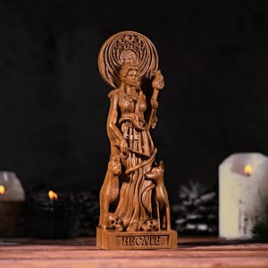May include: A brown wooden statue of the figure Hecate, featuring a circular halo, a torch, a snake, two dogs, and two skulls at the base. The name HECATE is carved into the rectangular base of the sculpture, which stands upright.