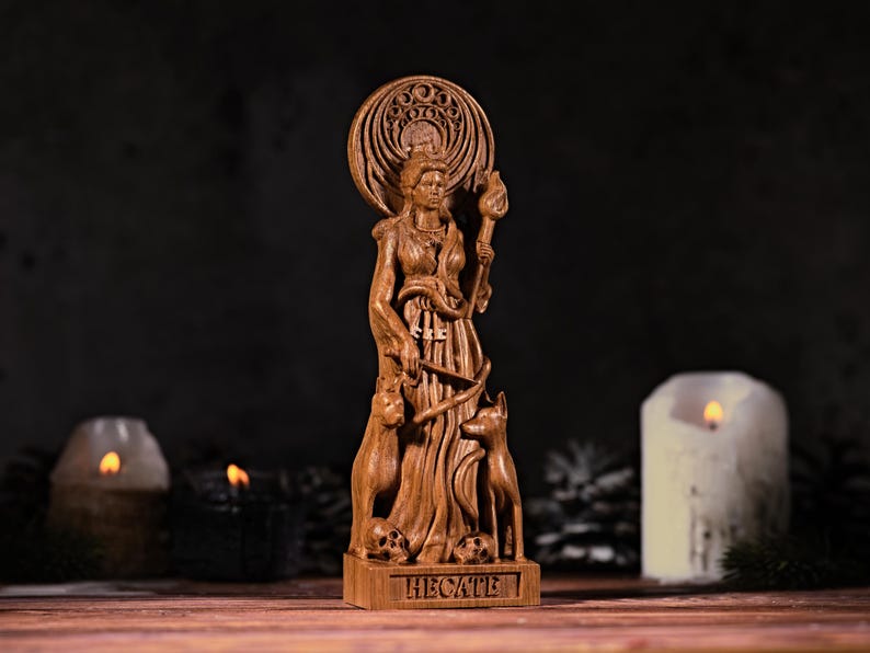 May include: A brown wooden statue of the figure Hecate, featuring a circular halo, a torch, a snake, two dogs, and two skulls at the base. The name HECATE is carved into the rectangular base of the sculpture, which stands upright.