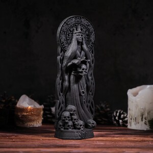 Puede incluir: Estatua gris oscura de una figura vestida con una t&uacute;nica, sosteniendo calaveras, con un halo y una corona. La base de la estatua est&aacute; rodeada de m&aacute;s calaveras. El fondo incluye velas y un ambiente oscuro, creando un tema g&oacute;tico o de fantas&iacute;a.