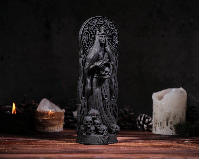 Puede incluir: Estatua gris oscura de una figura vestida con una t&uacute;nica, sosteniendo calaveras, con un halo y una corona. La base de la estatua est&aacute; rodeada de m&aacute;s calaveras. El fondo incluye velas y un ambiente oscuro, creando un tema g&oacute;tico o de fantas&iacute;a.