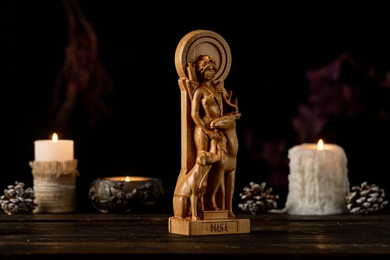 May include: A wooden statue of a woman with a deer and a dog. The statue is carved in a detailed style and has a rustic look. The woman is holding a bow and arrow. The statue is standing on a wooden base with the word "MARA" inscribed on it.