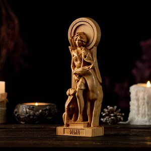 May include: A wooden statue of the Roman goddess Diana, depicted with a bow and arrow, standing with a deer and a dog. The statue is on a wooden base with the word "Diana" inscribed on it. The statue is surrounded by candles and pine cones.