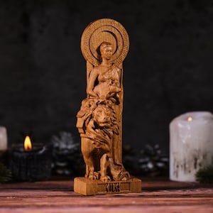 Op de afbeelding: Een houten beeld van een vrouw met een leeuw. Het beeld is gemaakt van lichtbruin hout en heeft ingewikkelde details. De vrouw heeft een halo en houdt een klein dier vast. De basis van het beeld heeft het woord "KIKK" erin gegraveerd.