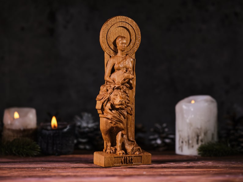 Op de afbeelding: Een houten beeld van een vrouw met een leeuw. Het beeld is gemaakt van lichtbruin hout en heeft ingewikkelde details. De vrouw heeft een halo en houdt een klein dier vast. De basis van het beeld heeft het woord "KIKK" erin gegraveerd.