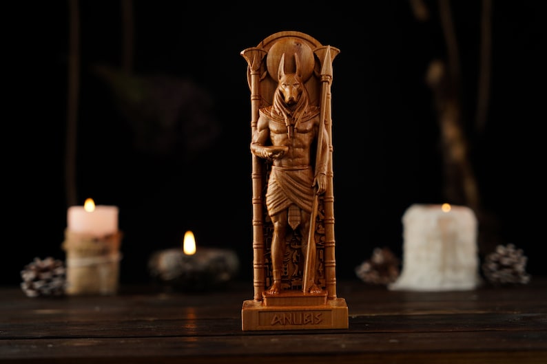 May include: A wooden Anubis statue, intricately carved with details of the Egyptian god. The statue is brown and stands upright, holding a staff. The word "ANUBIS" is carved on the base. The background is dark, with lit candles and pine cones.