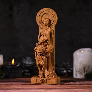 Op de afbeelding: Een houten beeld van een vrouw met een leeuw. De vrouw houdt een klein dier vast. Het beeld is lichtbruin en staat op een rechthoekige basis. Achter het hoofd van de vrouw bevindt zich een cirkelvormige halo.