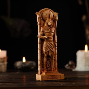 May include: A wooden Anubis statue, a deity from ancient Egyptian mythology, stands tall. The statue is carved in detail, depicting the jackal-headed god in a standing pose. The word "ANUBIS" is carved on the base. The statue is illuminated by candlelight.
