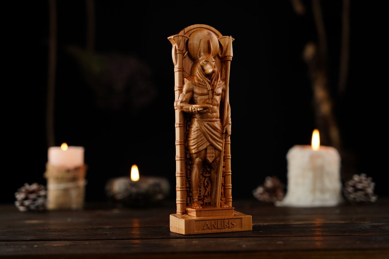 May include: A wooden Anubis statue, a deity from ancient Egyptian mythology, stands tall. The statue is carved in detail, depicting the jackal-headed god in a standing pose. The word "ANUBIS" is carved on the base. The statue is illuminated by candlelight.