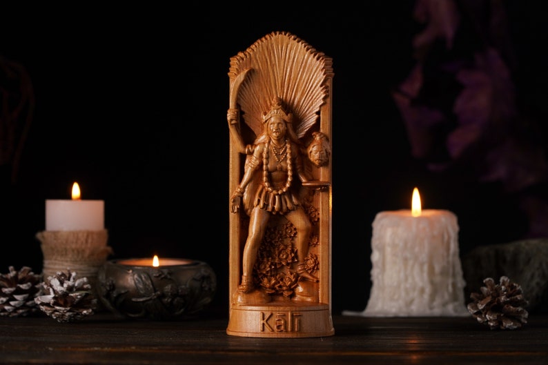 Pu&ograve; includere: Statua in legno della dea ind&ugrave; Kali, intagliata con dettagli. La figura tiene una spada e una testa mozzata. La statua &egrave; ambientata su uno sfondo scuro con candele accese e la parola "Kali" alla base.