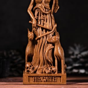 May include: A brown wooden statue of the goddess Hecate, featuring a central figure draped in robes, holding a sword and keys, flanked by two dogs and two skulls at the base. The rectangular base is engraved with the name HECATE in capital letters.
