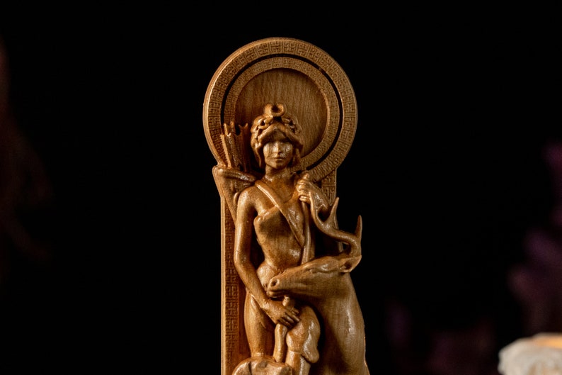 May include: A wooden carving of a woman with a deer, she is holding a bow and arrow. The carving is in a circular frame with a decorative border.