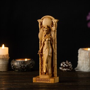 Op de afbeelding: Een houten beeld van Nephthys, een Egyptische godin, is afgebeeld. Het beeld is gedetailleerd gesneden en toont haar kleding en hoofdtooi. De basis van het beeld is gegraveerd met de naam "Nephthys". De achtergrond bevat kaarsen en decoratieve elementen.