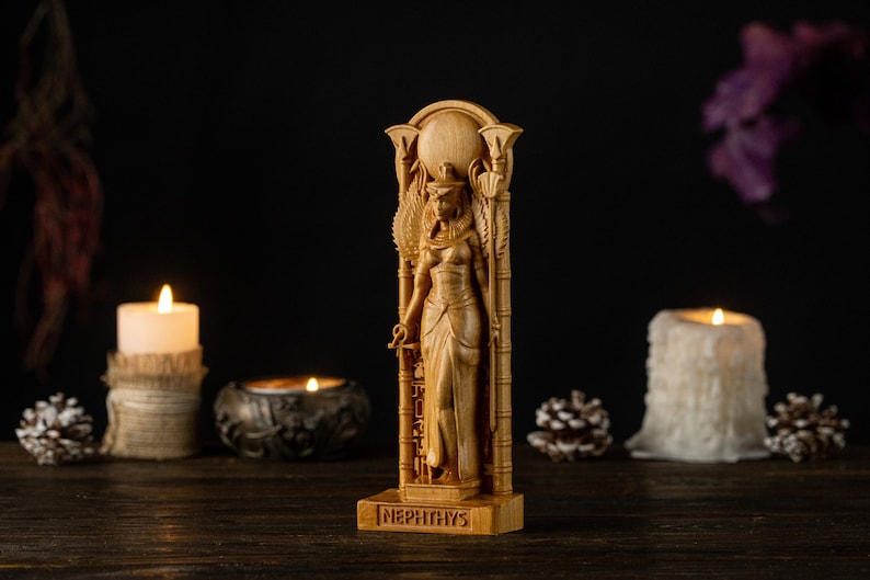 Op de afbeelding: Een houten beeld van Nephthys, een Egyptische godin, is afgebeeld. Het beeld is gedetailleerd gesneden en toont haar kleding en hoofdtooi. De basis van het beeld is gegraveerd met de naam "Nephthys". De achtergrond bevat kaarsen en decoratieve elementen.