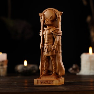 Pode incluir: Escultura de madeira detalhada de uma figura com cabe&ccedil;a de lobo, usando armadura e capa, segurando um bast&atilde;o. A base da escultura tem a palavra "WAPWAWET" esculpida. A escultura est&aacute; em um fundo escuro.