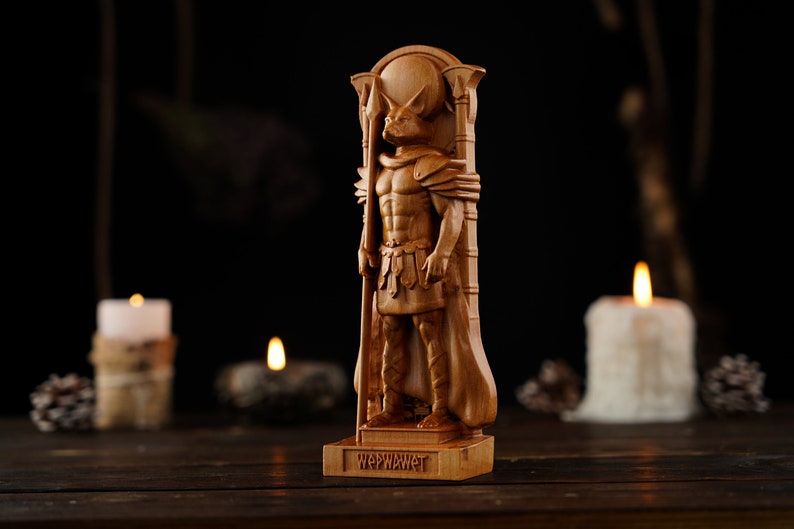 Pode incluir: Escultura de madeira detalhada de uma figura com cabe&ccedil;a de lobo, usando armadura e capa, segurando um bast&atilde;o. A base da escultura tem a palavra "WAPWAWET" esculpida. A escultura est&aacute; em um fundo escuro.