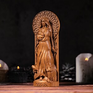 May include: A wooden statue of a woman holding a bundle, with the word "OSTARA" at the base. The statue is intricately carved with a detailed design, and is set against a dark background with lit candles.